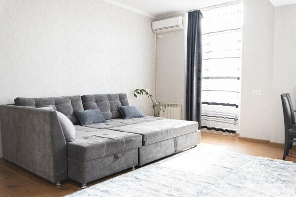 A grey sectional sofa with cushions, a white radiator, a black curtain, and a plant in a room with light-colored walls and wooden flooring.
