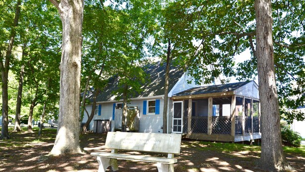 A wooden bench in a shaded area with a house in the background.
