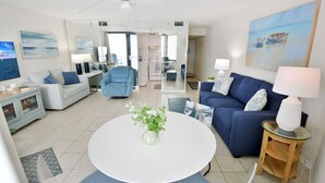 A modern living room with a white round table, a blue sofa, and a blue armchair. There is a kitchen area in the background with a refrigerator and a microwave. - Beach Front Condo with Endless Gulf Views!   (Panama City Beach)