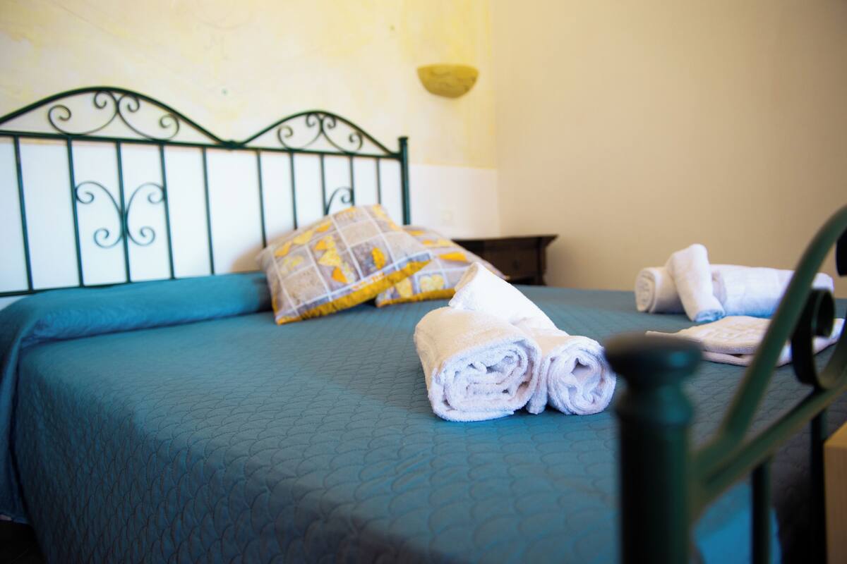 Ein Bett mit einer blauen Steppdecke, einem gelb-weißen Kopfkissen und zusammengerollten Handtüchern daneben.