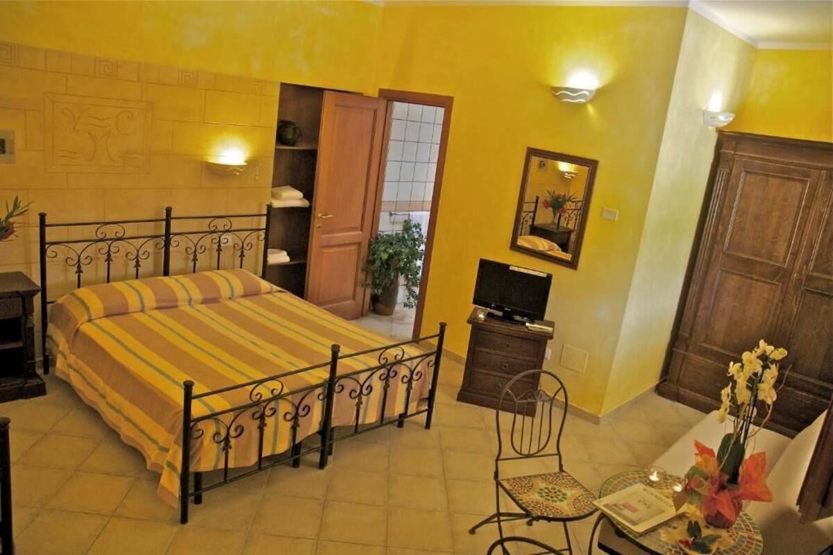 Ein Schlafzimmer mit einem Bett, einem Fernseher, einem Sessel, einem kleinen Tisch mit einer Vase und einer Tür zu einem weiteren Raum.