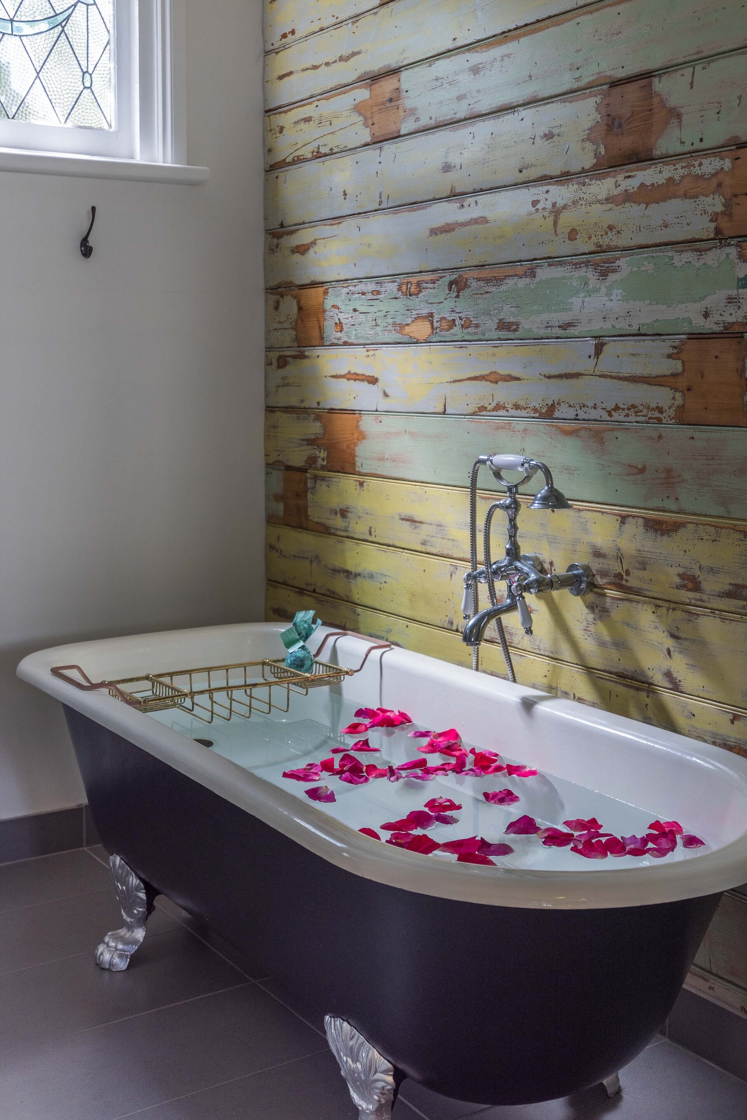 Una bañera de patas de león con pétalos de rosa en el agua, un tendedero metálico y una pared de madera rústica.