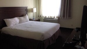 Ein Hotelzimmer mit einem großen Bett, einem Nachttisch mit Lampe, einem Fernseher und einem Fenster mit Vorhängen.