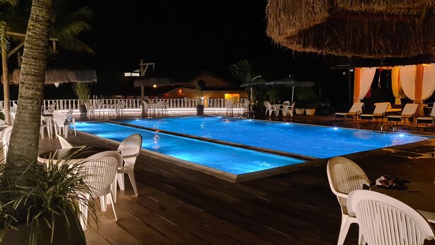 Un área de piscina nocturna con sillones de descanso y una estructura de techo de paja.