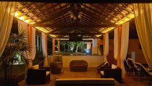 Un espacio de estar al aire libre cubierto, con vista a una piscina y palmeras.