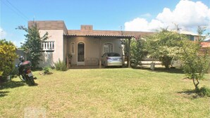 A single-story house with a car parked in the driveway, surrounded by a small lawn and a few trees. - Casa Pátio Grande / 8 people - Praia do Moçambique (Florianópolis)