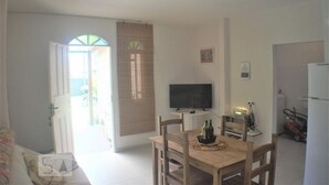 A dining area with a wooden table and chairs, a television on the wall, and a kitchen area with a refrigerator and a lawnmower. - Casa Pátio Grande / 8 people - Praia do Moçambique (Florianópolis)