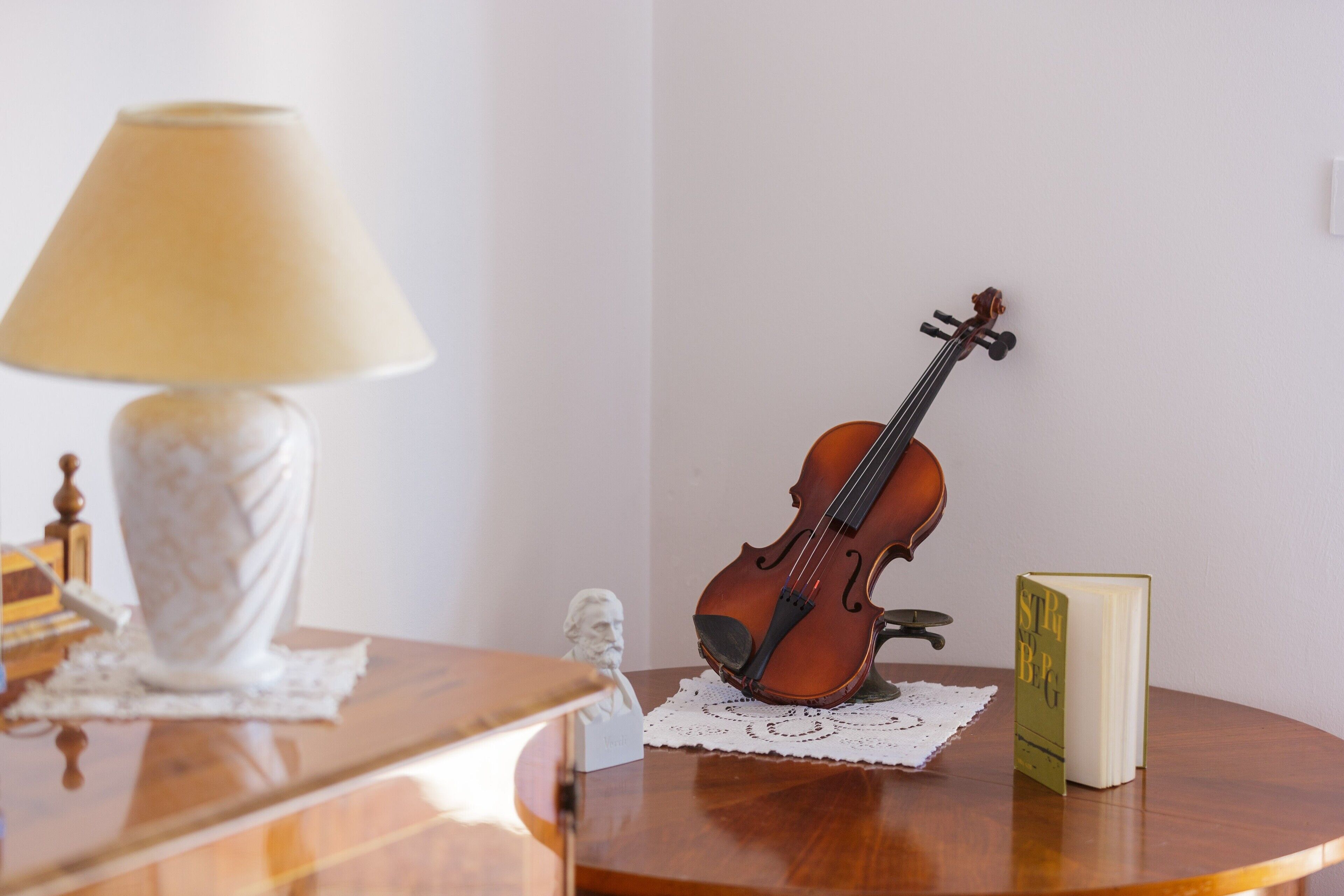 Un violín sobre una mesa de madera con un mantel de encaje, una lámpara y un libro.