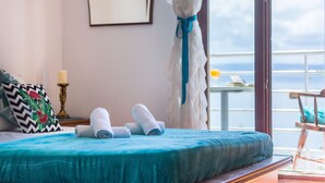 Un lit recouvert d’une couvre-lit turquoise, agrémenté de serviettes blanches et d’un coussin décoratif. Un rideau est retenu sur le côté, dévoilant un balcon avec une balustrade et une table.