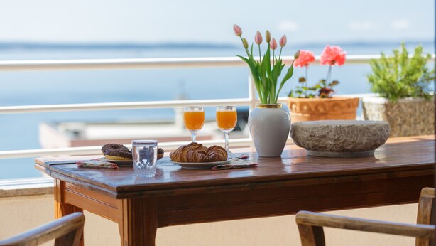 Une table en bois avec des croissants, du jus d’orange et un vase de fleurs, le tout sur un balcon donnant sur la mer.