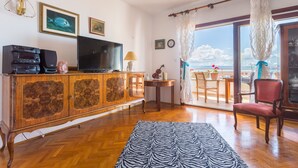 Un salon avec un meuble en bois, un téléviseur à écran plat, un tapis à motifs zébrés, un fauteuil rouge et une vue sur la mer par une porte vitrée.