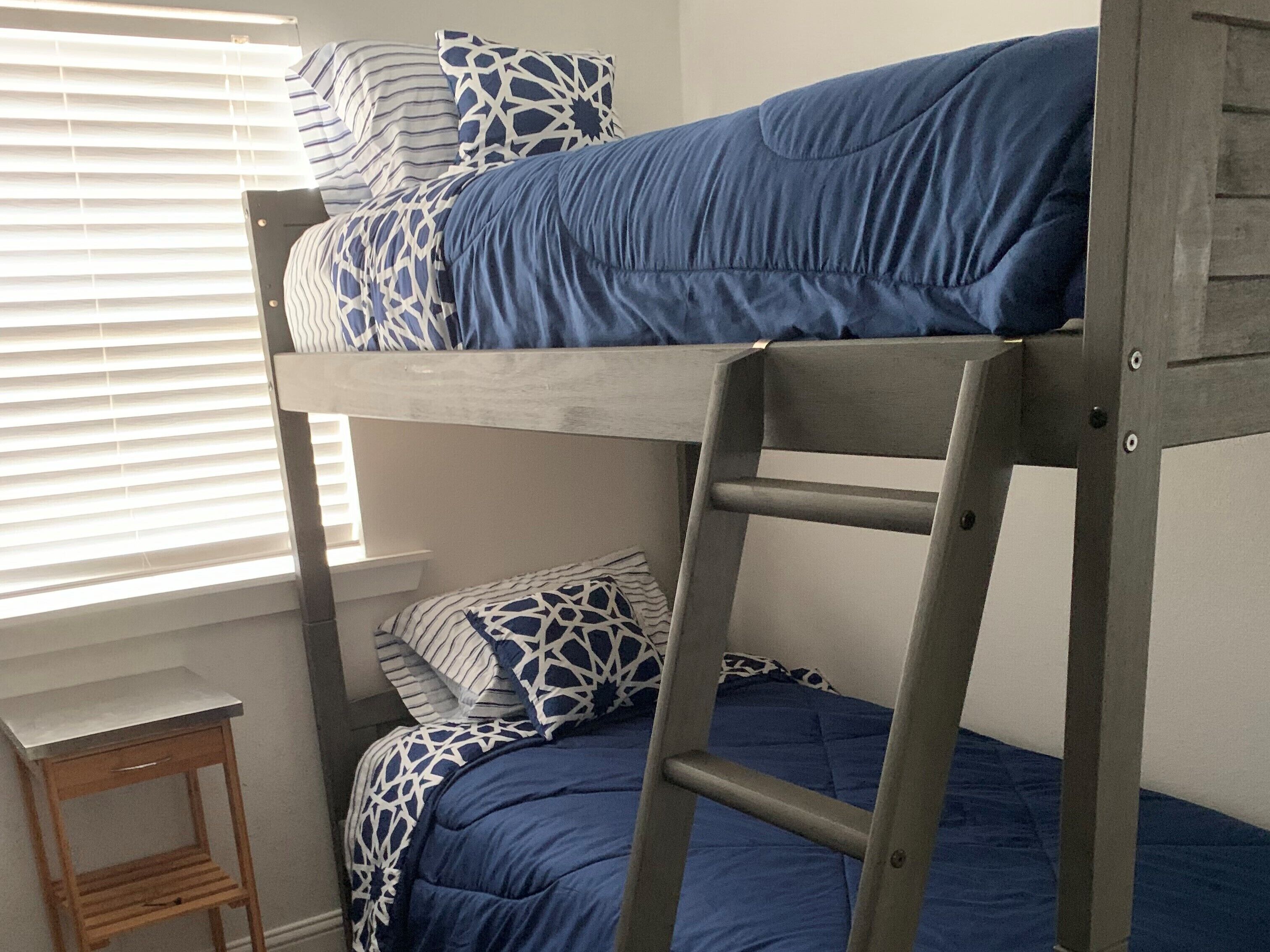 Una cama litera con ropa de cama azul y almohadas estampadas, una escalera de madera y un taburete pequeño de madera.