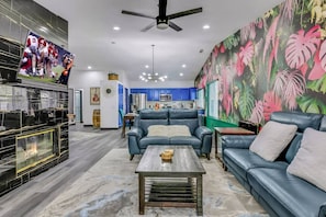 A living room with a fireplace, a blue sofa set, a coffee table, and a large tropical-themed mural. - Casa Del Sol: #259070 Pool, Spa, Game Room, Court (La Quinta)