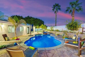 A swimming pool with lounge chairs and umbrellas, surrounded by a landscaped garden and a building with windows. - Casa Del Sol: #259070 Pool, Spa, Game Room, Court (La Quinta)