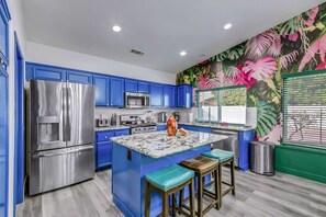A modern kitchen with blue cabinets, stainless steel appliances, a central island with stools, and a vibrant tropical mural. - Casa Del Sol: #259070 Pool, Spa, Game Room, Court (La Quinta)