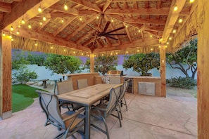 An outdoor patio with a wooden table and chairs, a grill, and string lights. - Casa Del Sol: #259070 Pool, Spa, Game Room, Court (La Quinta)
