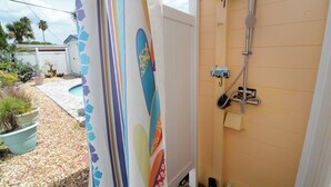 Un piccolo box doccia chiuso con un tubo flessibile e un soffione, una pianta in vaso e un tavolozza da surf appoggiata a una parete.