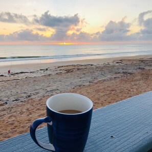 Una taza azul sobre una cubierta de madera con vista a una playa al atardecer.