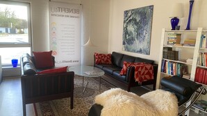 En moderne stue med sofa, lænestol og sk footmop, reol og et stort abstrakt maleri.