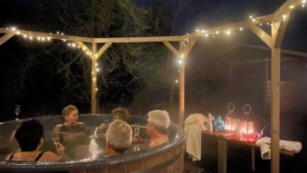 Et boblebad med mennesker omkring, en træpergola med lyskæder, og et bord med stearinlys.
