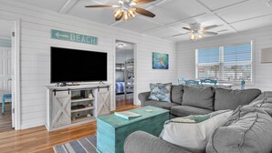 A living room with a television, a couch, a coffee table, and a sign indicating a beach direction. - Salty But Sweet: Renovated Oceanfront Home on Oak Island with Great Views (Oak Island)