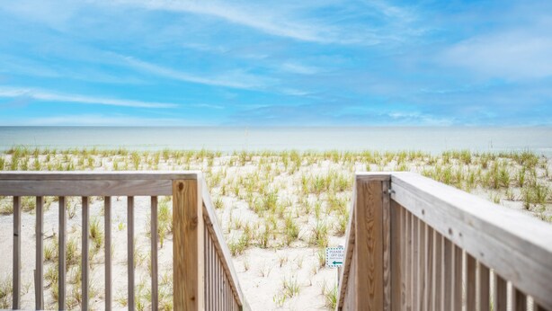 A wooden deck leading to a beach with sand and grass, and a sign indicating a direction. - Salty But Sweet: Renovated Oceanfront Home on Oak Island with Great Views (Oak Island)