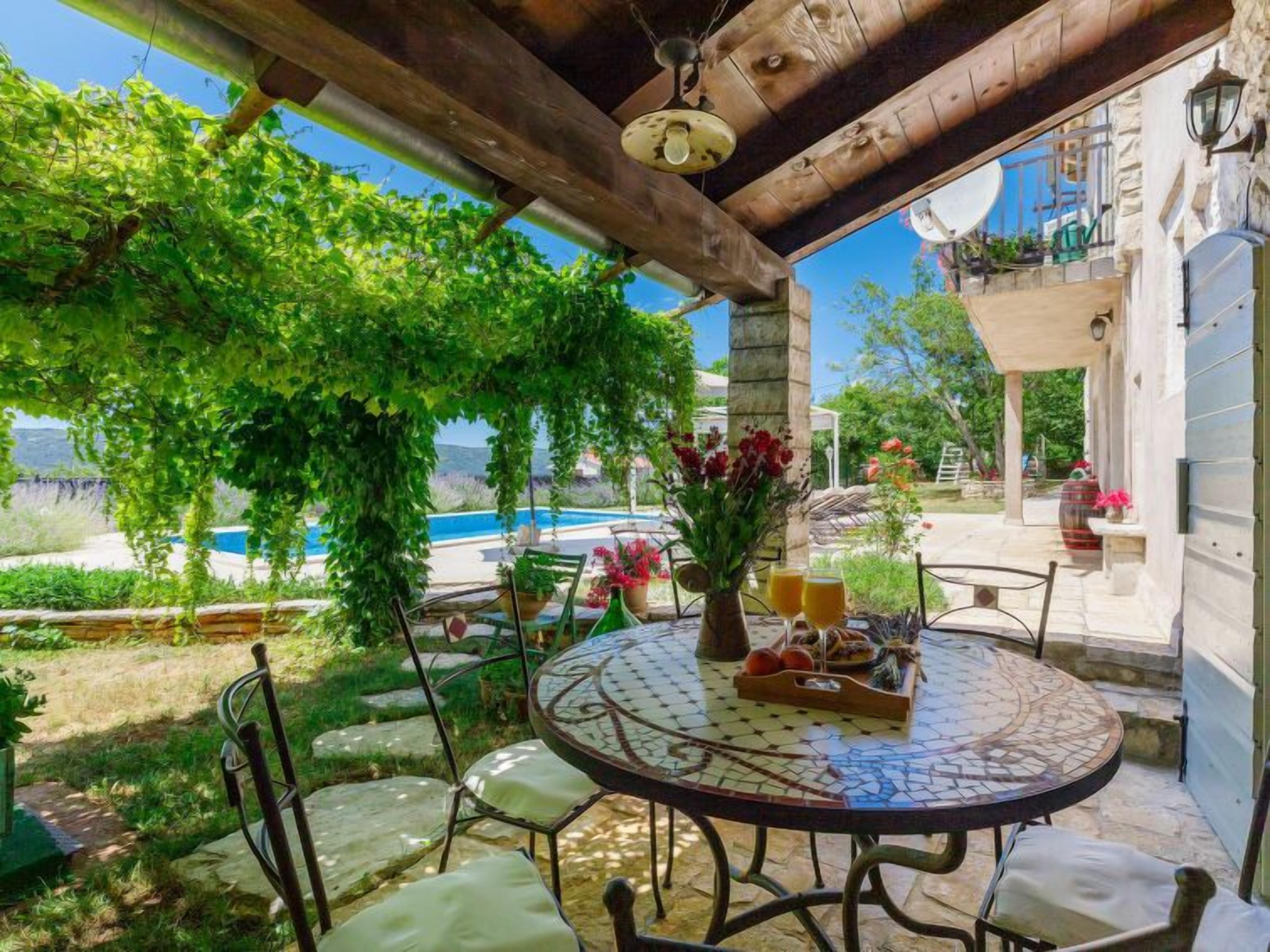 Une terrasse extérieure couverte, agrémentée d’une table à motifs de mosaïque, de chaises et offrant une vue sur une piscine et la verdure environnante.