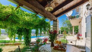 Une terrasse extérieure couverte, agrémentée d’une table à motifs de mosaïque, de chaises et offrant une vue sur une piscine et la verdure environnante.