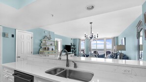 A kitchen with a large island, a double sink, and a dishwasher. There is a chandelier above the island and a view of the living room through the window.