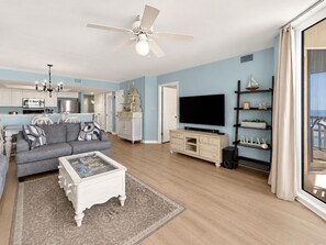 A living room with a grey sofa, a coffee table, and a TV. There is a ceiling fan, a chandelier, and a bookshelf with decorative items.