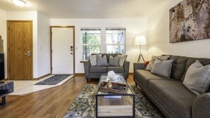 Een woonkamer met een bank, een bijzettafeltje en een vloerkleed. Er is een deur en ramen te zien op de achtergrond.