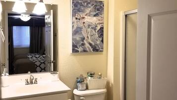 Un baño con un lavamanos blanco, inodoro y ducha. Hay un cuadro en la pared y una ventana con cortinas.