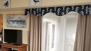Un dormitorio con cama, un televisor sobre un mueble de madera, una ventana con cortinas y vistas a una terraza exterior.