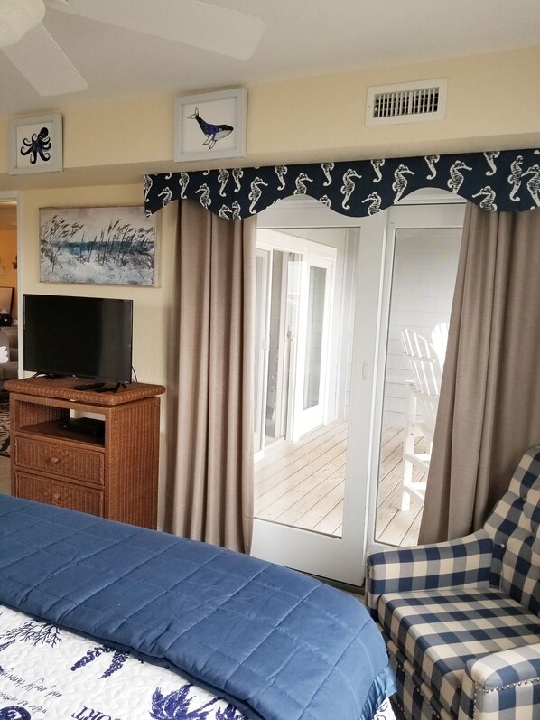 A bedroom with a bed, a television on a wooden stand, a window with curtains, and a view of an outdoor deck. - OCEAN/SOUND VIEW!OCEAN BOARDWALK!  BEACH EQUIP! BABY EQUIP! GUEST FAVORITE! (Rodanthe)