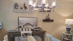 Un comedor con una mesa de vidrio, sillas blancas, una araña, obras de arte enmarcadas y un cuenco decorativo.