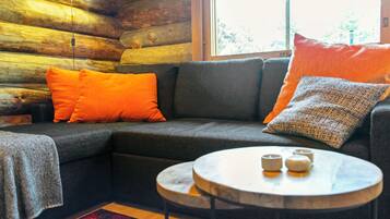 L’intérieur d’un chalet confortable, avec une cheminée, un canapé d’angle, une table basse ronde et une fenêtre donnant sur la forêt.