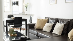 Una sala moderna con un sofá de cuero negro, una mesa de centro de vidrio y un comedor al fondo.