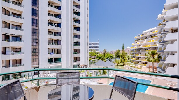 Un balcón con una mesa y sillas de vidrio, con vista a una piscina y edificios cercanos.