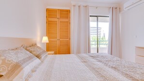 A bedroom with a large bed, a wooden wardrobe, a window with curtains, and a balcony view. - Taalia I - Vilamoura (Vilamoura)