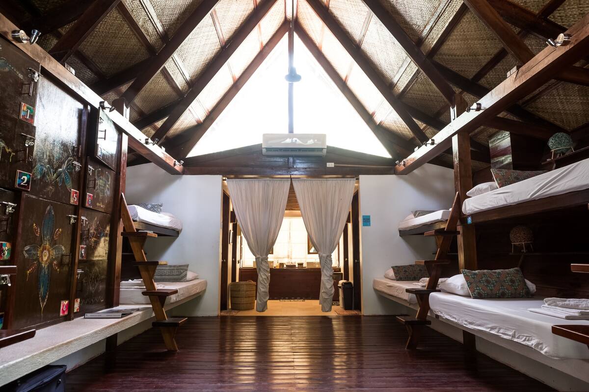 A dormitory room with bunk beds, wooden beams, a ceiling fan, and a window with curtains.