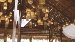 Een binnenzittplaats met houten tafels en banken, hangende rieten lampen en een rieten dakconstructie.