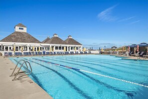 A swimming pool with clear lane dividers, a lounge area with blue chairs, and a building with a gabled roof.