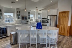 A modern kitchen with white cabinetry, stainless steel appliances, and a central island with bar stools. - Ocean Hideaway | Oceanfront | Community Pool (Nags Head)