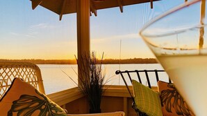 A wicker chair with a cushion, a glass of a beverage, and a view of a lake at sunset.