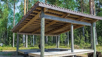 Un padiglione in legno con tetto ondulato, sorretto da pali di legno, immerso in una zona boschiva.