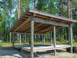 A wooden pavilion with a corrugated roof, supported by wooden posts, set in a forested area.