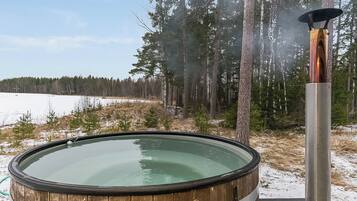 A wooden hot tub with a metal heater, surrounded by a snowy landscape and trees.