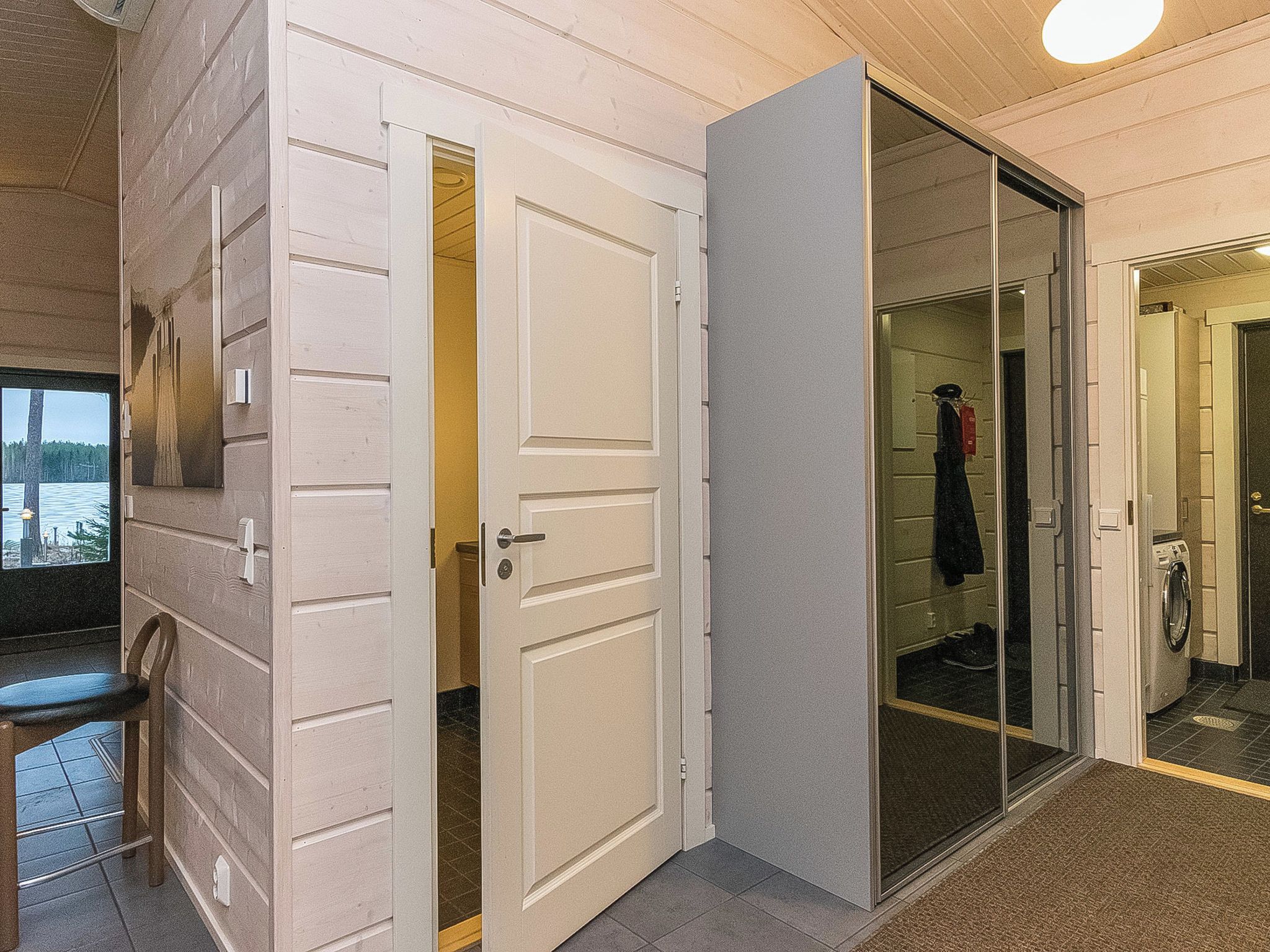 A hallway with a door leading to a bathroom, a chair, and a view of the outside.