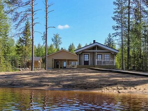 A cabin with a front porch and a small attached shed, surrounded by trees and a body of water.