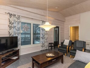 A modern living room with a flat-screen TV, a wooden coffee table, two armchairs, a sofa, and a large window with patterned curtains. - Villa mari by Interhome (Pertunmaa)